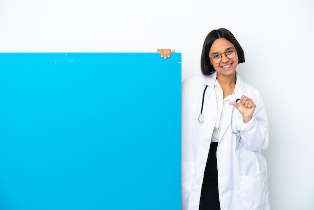 Mulher jovem médica mestiça com um grande cartaz isolado no fundo branco apontando para o lado para apresentar um produto