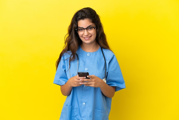 Mulher jovem médico cirurgião isolada em fundo amarelo enviando uma mensagem com o celular