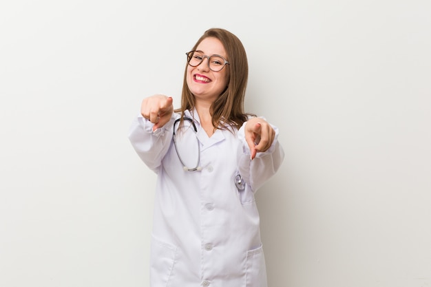 Mulher jovem médico contra um sorriso alegre de parede branca, apontando para a frente.