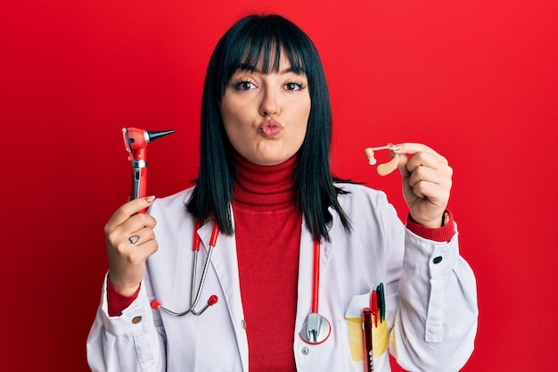 Mulher jovem médico hispânico segurando fone de ouvido de surdez e otoscópio olhando para a câmera mandando um beijo sendo adorável e sexy. expressão de amor.