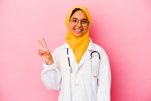 Mulher jovem médico muçulmana isolada na parede rosa, mostrando o sinal da vitória e sorrindo amplamente.