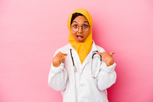 Mulher jovem médico muçulmana isolada na parede rosa surpresa apontando com o dedo, sorrindo amplamente.