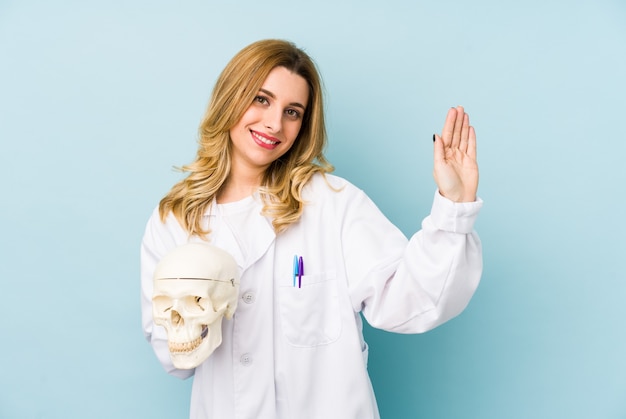 Mulher jovem médico segurando uma caveira isolada sorrindo alegre mostrando o número cinco com os dedos.