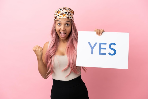 Mulher jovem mestiça com cabelo rosa isolada em um fundo rosa segurando um cartaz com o texto SIM