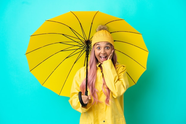 Mulher jovem mestiça com casaco à prova de chuva e guarda-chuva com expressão facial surpresa e chocada