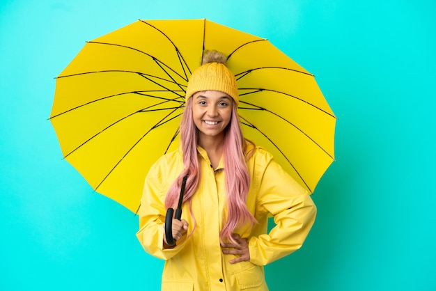 Mulher jovem mestiça com casaco à prova de chuva e guarda-chuva posando com os braços na cintura e sorrindo
