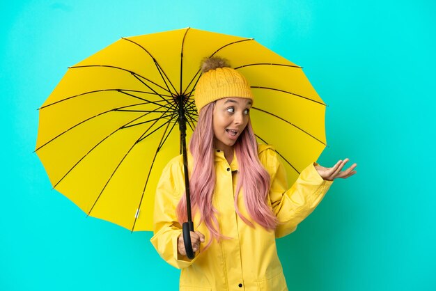 Mulher jovem mestiça com casaco impermeável e guarda-chuva com expressão surpresa enquanto olha para o lado