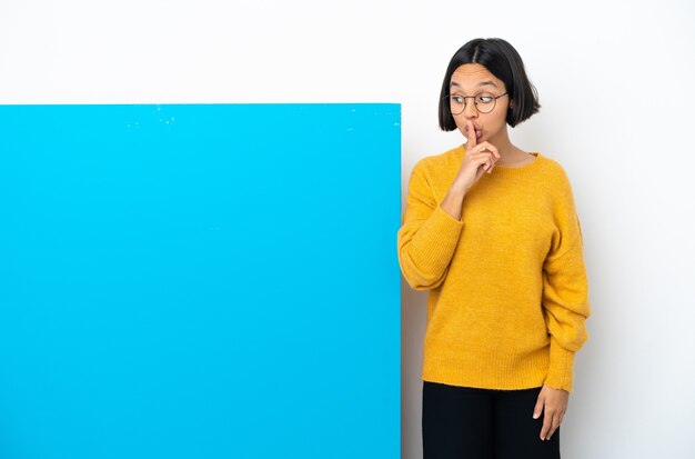 Mulher jovem mestiça com um grande cartaz azul isolado no fundo branco mostrando um sinal de silêncio gesto colocando o dedo na boca