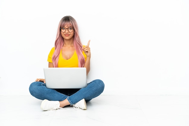 Mulher jovem mestiça com um laptop de cabelo rosa sentada no chão isolado no fundo branco apontando com o dedo indicador uma ótima ideia