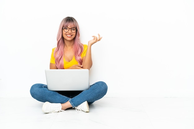 Mulher jovem mestiça com um laptop de cabelo rosa sentada no chão, isolado no fundo branco, estendendo as mãos para o lado para convidar para vir