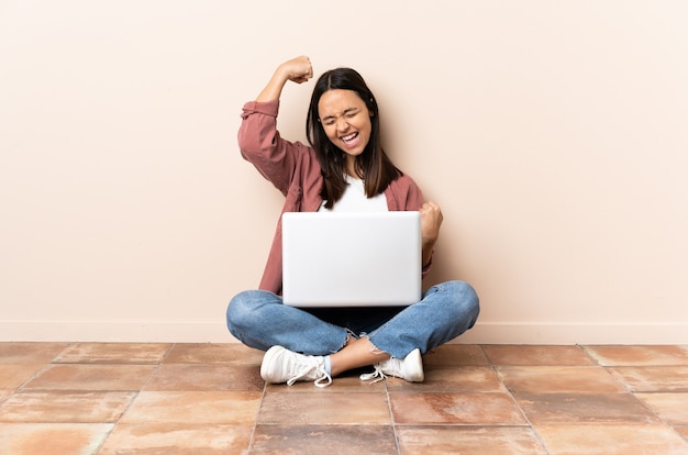 Mulher jovem mestiça com um laptop sentada no chão comemorando uma vitória