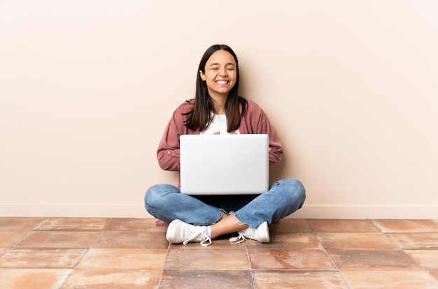 Mulher jovem mestiça com um laptop sentada no chão e sorrindo muito