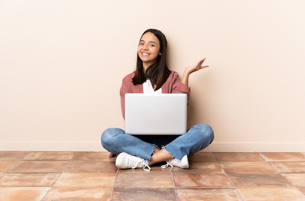 Mulher jovem mestiça com um laptop sentada no chão estendendo as mãos para o lado para convidar para vir