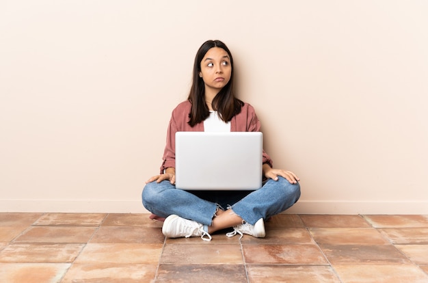 Mulher jovem mestiça com um laptop sentada no chão fazendo gestos de dúvida olhando para o lado