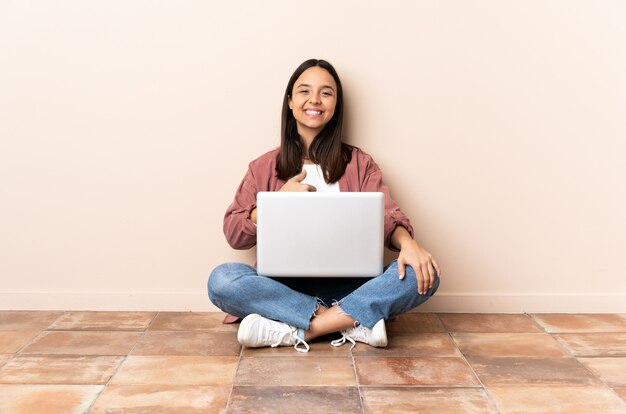 Mulher jovem mestiça com um laptop sentada no chão fazendo um gesto de polegar para cima