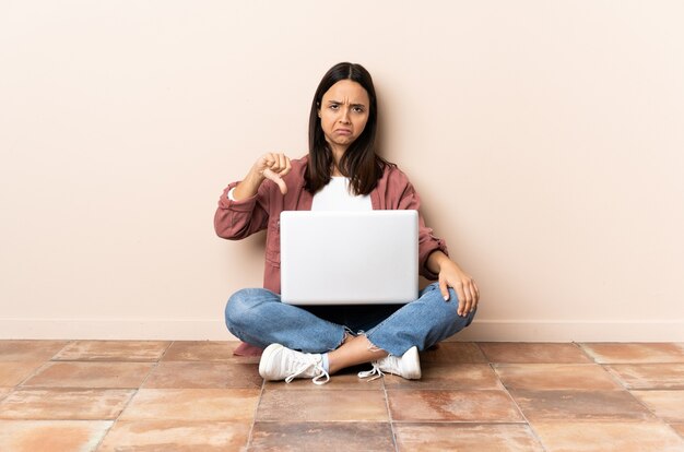 Mulher jovem mestiça com um laptop sentada no chão mostrando o polegar para baixo com expressão negativa