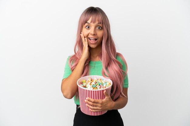 Mulher jovem mestiça de cabelo rosa comendo pipoca isolada no fundo branco com expressão facial surpresa e chocada