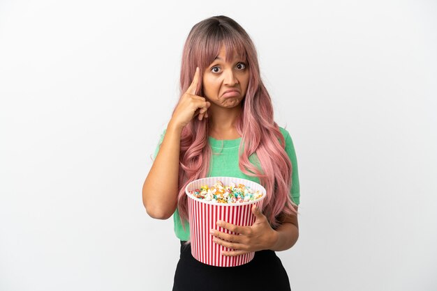 Foto mulher jovem mestiça de cabelo rosa comendo pipoca isolada no fundo branco pensando em uma ideia