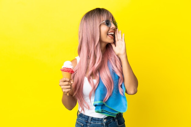 Mulher jovem mestiça de cabelo rosa segurando um sorvete isolado em um fundo amarelo e gritando com a boca bem aberta para o lado