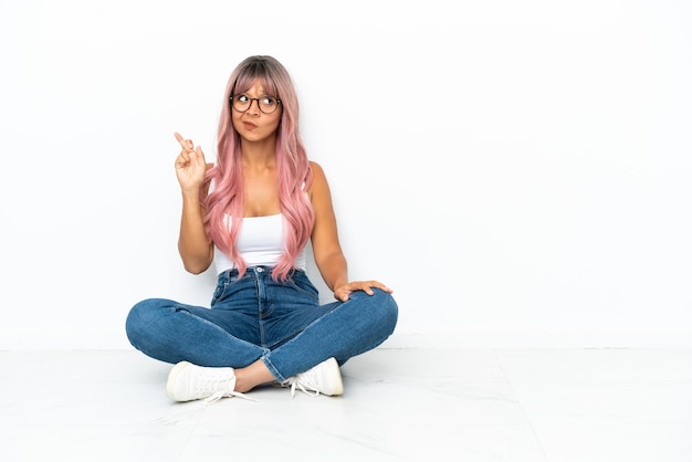 Mulher jovem, mestiça, de cabelo rosa, sentada no chão, isolada no fundo branco, com os dedos se cruzando e desejando o melhor