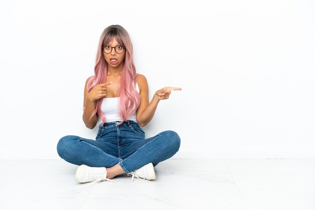 Mulher jovem mestiça de cabelo rosa sentada no chão isolada no fundo branco surpresa e apontando para o lado