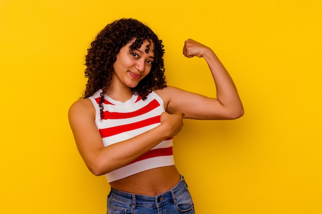 Mulher jovem mestiça isolada em fundo amarelo mostrando gesto de força com os braços, símbolo do poder feminino