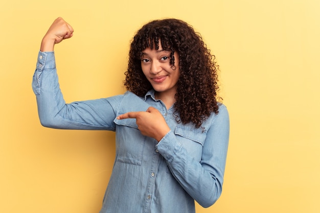 Foto mulher jovem mestiça isolada em fundo amarelo mostrando gesto de força com os braços, símbolo do poder feminino