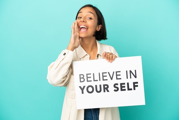 Mulher jovem mestiça isolada em um fundo azul segurando um cartaz com o texto acredite em si mesmo e gritando