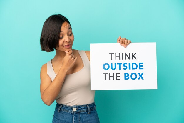 Mulher jovem mestiça isolada em um fundo azul segurando um cartaz com o texto pense fora da caixa e pensando