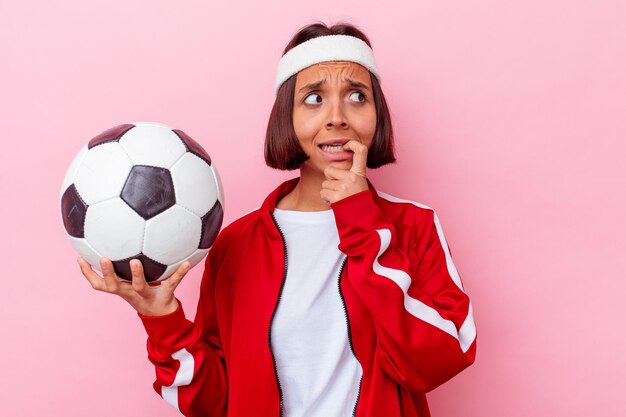 Mulher jovem mestiça jogando futebol isolada na parede rosa, roendo as unhas, nervosa e muito ansiosa.