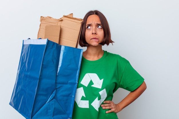 Mulher jovem mestiça reciclando papelão isolado na parede branca confusa, sente-se em dúvida e insegura.