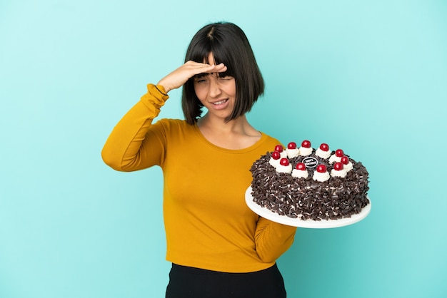 Mulher jovem mestiça segurando bolo de aniversário olhando para longe com a mão para ver algo