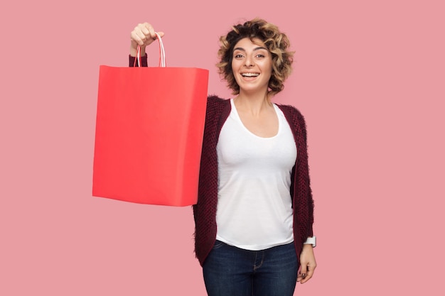 Foto mulher jovem moderna satisfeita com camisa azul com penteado cacheado em pé e mostrando sacolas de compras e sorriso cheio de dentes, olhando para a câmera. foto de estúdio, fundo rosa, isolado, interior, espaço de cópia