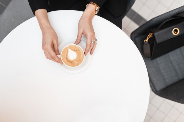 Mulher jovem moderna se senta em uma mesa branca vintage em um café e segura uma xícara de cappuccino delicioso quente. vista superior em cima da mesa com café nas mãos femininas.