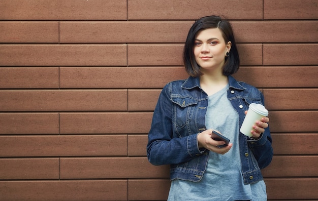 Mulher jovem morena atraente com túneis nas orelhas em uma jaqueta jeans com smartphone em frente a parede de tijolos, desfrutando de seu café quente.