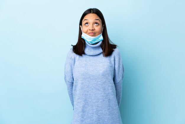 Mulher jovem morena de raça mista, protegendo com uma máscara e luvas sobre parede azul e olhando para cima