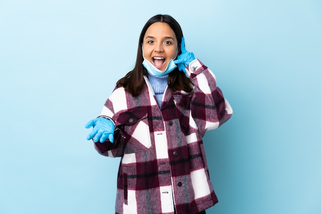 Mulher jovem morena de raça mista, protegendo com uma máscara e luvas sobre parede azul, fazendo gesto de telefone e duvidando