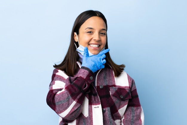 Mulher jovem morena de raça mista, protegendo com uma máscara e luvas sobre parede azul feliz e sorridente