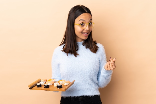 Foto mulher jovem morena de raça mista, segurando o sushi sobre parede isolada, fazendo gesto de dinheiro