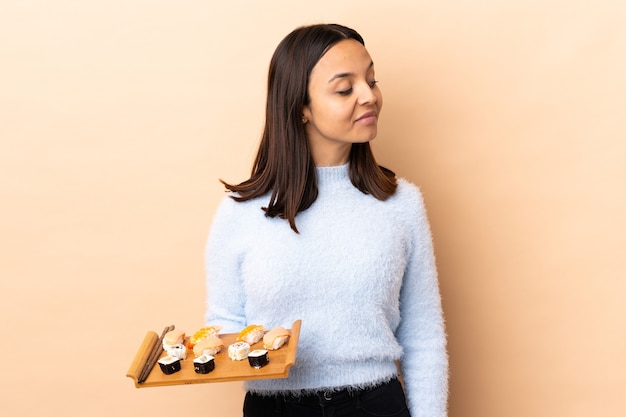 Foto mulher jovem morena de raça mista segurando sushi sobre uma parede isolada com dúvidas enquanto olha de lado