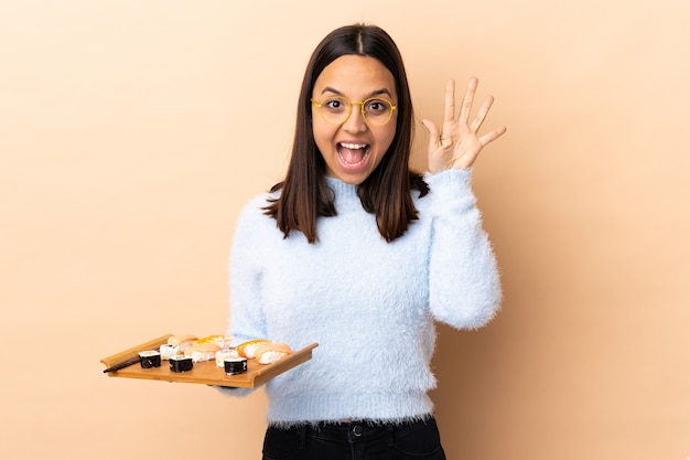 Mulher jovem morena de raça mista segurando sushi sobre uma parede isolada, contando cinco com os dedos