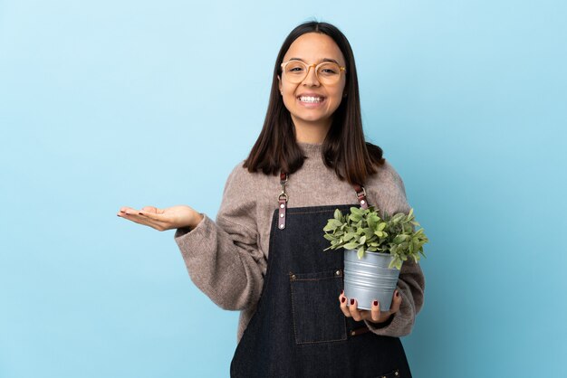 Mulher jovem morena de raça mista, segurando uma planta sobre fundo azul isolado, segurando copyspace imaginário na palma da mão para inserir um anúncio