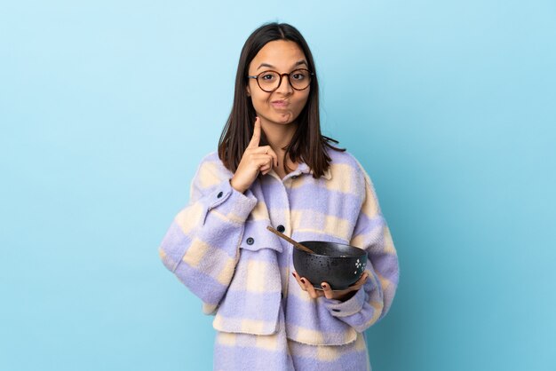 Mulher jovem morena de raça mista, segurando uma tigela cheia de macarrão sobre parede azul e pensamento.