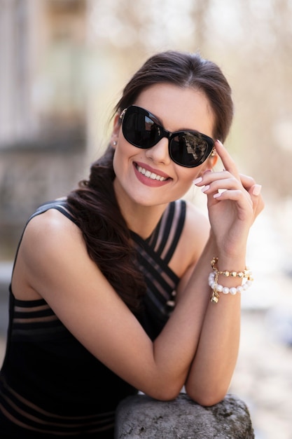 Mulher jovem morena elegantemente linda alegre, num elegante vestido preto e óculos escuros, posando do lado de fora, tocando as mãos juntas na varanda.