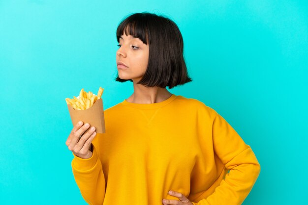 Mulher jovem morena segurando batatas fritas sobre um fundo azul isolado, olhando para o lado