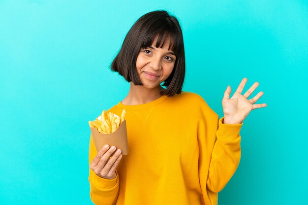 Mulher jovem morena segurando batatas fritas sobre uma parede azul isolada e saudando com a mão com uma expressão feliz