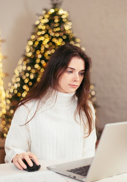 Mulher jovem morena trabalha em casa remotamente, atrás de um laptop e com documentos no fundo de uma árvore de Natal. ambiente festivo e trabalho em casa. 2021