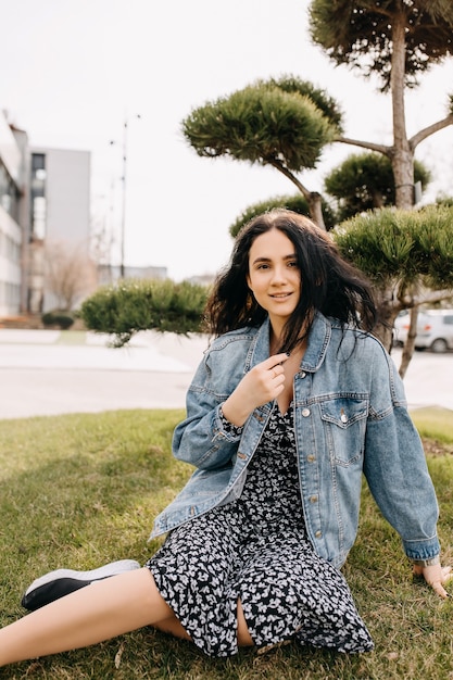 Mulher jovem morena vestindo uma jaqueta jeans, sentada na grama verde em um parque