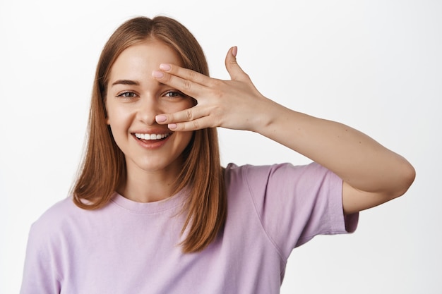 Foto mulher jovem mostrando seu rosto limpo, pele facial sem maquiagem, aparência feminina natural, dedos abertos perto dos olhos e sorrindo feliz, encostada em uma parede branca