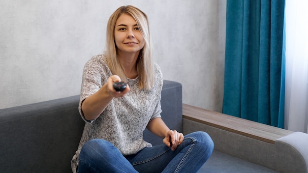 Mulher jovem muda os canais de televisão com o controle remoto. Ela sorri e olha para a câmera